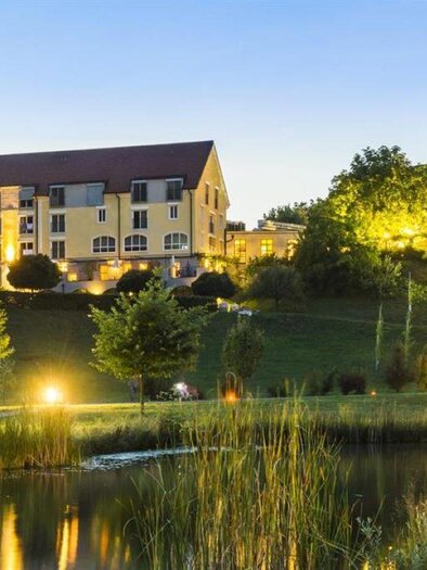 Ein ruhiges Hotelgebäude liegt am Ufer eines Teichs, umgeben von Bäumen und grüner Landschaft. Die Abendbeleuchtung verleiht der Szene eine einladende Atmosphäre. | © Hotel-Restaurant Staribacher
