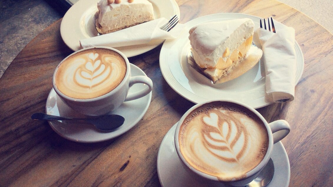 Zwei Tassen Kaffee mit Latte-Art auf einem Holz-Tisch neben zwei Stück Kuchen. Die Atmosphäre ist gemütlich und einladend. | © Shutterstock_Mutita Narkmuang
