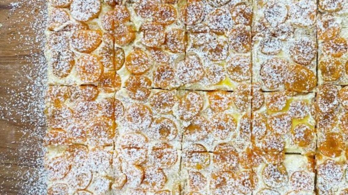 Ein quadratisches Dessert mit fruchtigem Belag und Puderzucker. Es ist in kleine Stücke geschnitten und liegt auf einem Holzbrett. | © Bäckerei-Konditorei Hubmann Josef GmbH