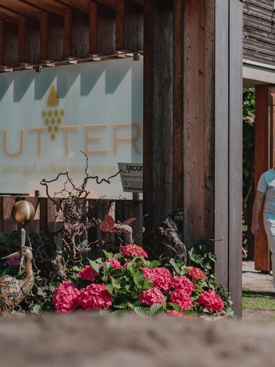 Ein Paar spaziert Hand in Hand am Eingangsbereich des Weinguts Hutter in Feldbach vorbei. Das Holzgebäude mit Weingut-Schild und blühenden Hortensien liegt im Thermen- & Vulkanland in der Steiermark. | © Thermen- & Vulkanland