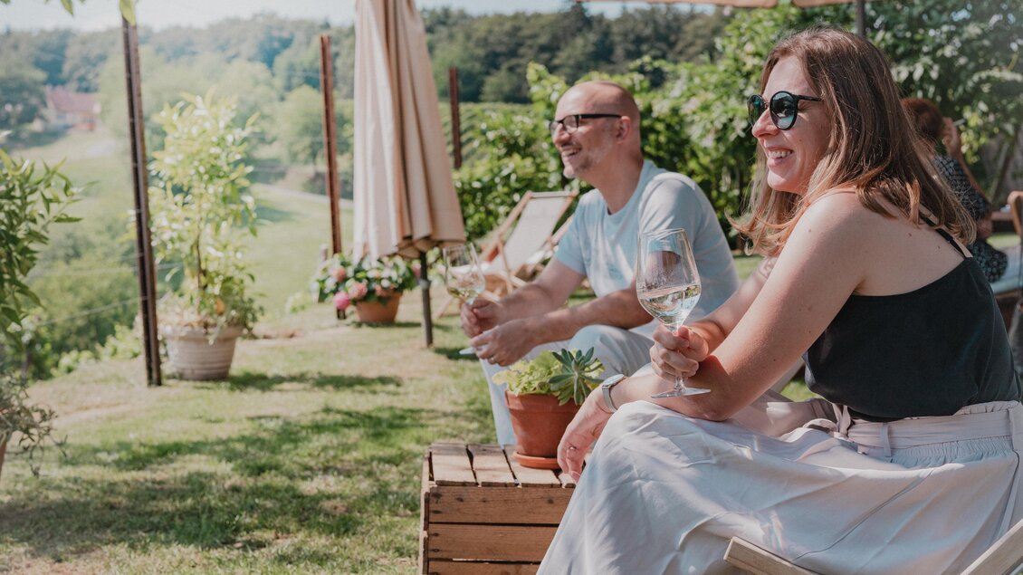 Zwei Gäste genießen ein Glas Weißwein im Garten des Weinguts Hutter in Feldbach mit Blick auf die grüne Hügellandschaft im Thermen- & Vulkanland in der Steiermark. | © Thermen- & Vulkanland