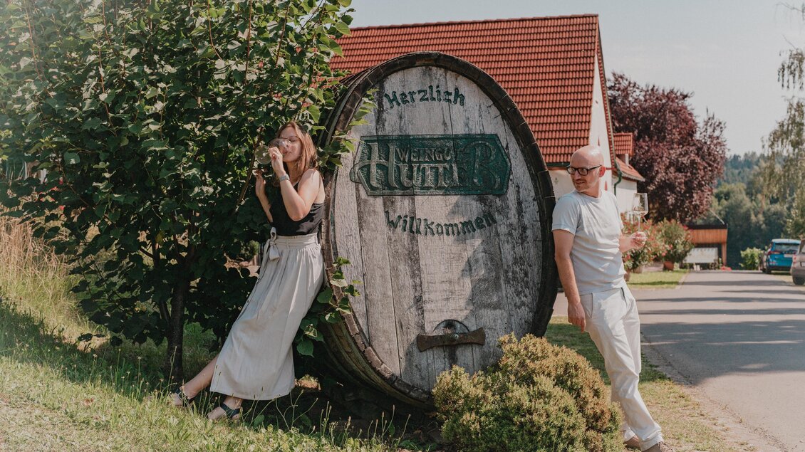 Zwei Gäste genießen Weißwein neben einem großen historischen Holzfass mit der Aufschrift Weingut Hutter in Feldbach. Ein Einblick in die Weinkultur des Thermen- & Vulkanlands. | © Thermen- & Vulkanland