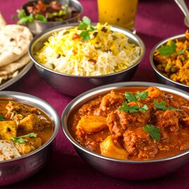Ein Tablett mit verschiedenen indischen Gerichten: Zwei Schalen mit Curry, darunter eines mit Hühnchen und eines mit Lamm, dazu Naan-Brot, duftender Reis und eine Portion Gemüse-Curry, garniert mit frischem Koriander. | © Firefly_Sarah Herzog