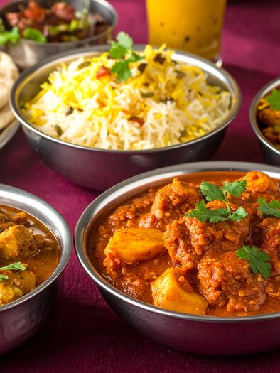 Ein Tablett mit verschiedenen indischen Gerichten: Zwei Schalen mit Curry, darunter eines mit Hühnchen und eines mit Lamm, dazu Naan-Brot, duftender Reis und eine Portion Gemüse-Curry, garniert mit frischem Koriander. | © Firefly_Sarah Herzog