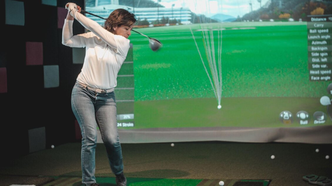 Eine Person spielt Golf in einem Indoor-Golfstudio. Im Hintergrund ist ein Bildschirm mit Golfstatistiken und einer virtuellen Golfbahn zu sehen. | © Indoor Golf Studio