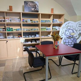 Ein Empfangsbereich mit einem Tisch und Stühlen. Im Hintergrund sind Regale mit Broschüren und ein freundliches Maskottchen zu sehen. | © TVB Südsteiermark