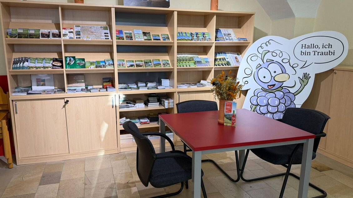 Ein Empfangsbereich mit einem Tisch und Stühlen. Im Hintergrund sind Regale mit Broschüren und ein freundliches Maskottchen zu sehen. | © TVB Südsteiermark
