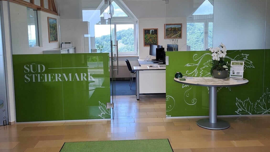 Ein modernes Büro mit grünen Wänden und einem Empfangstisch. Der Boden ist mit einem Teppich mit der Aufschrift "SÜDSTEIERMARK" ausgestattet. | © TV Südsteiermark