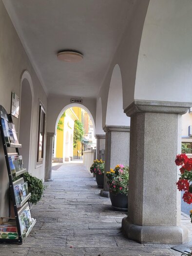 Ein schmaler Durchgang mit Steinboden und hohen Bögen. An den Wänden stehen Zeitungsständer und blühende Pflanzen sorgen für Farbe. | © TV Südsteiermark - Irene Löschnig
