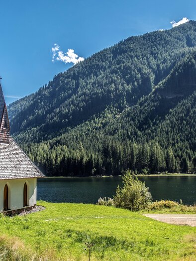 A small church stands by the shore of a clear lake, surrounded by high mountains and dense forests. The sky is blue with a few clouds. | © Anita Fössl