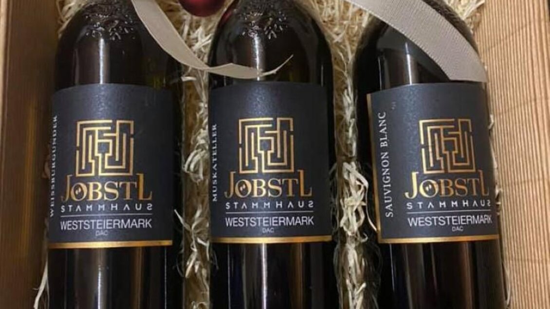 Drei Flaschen Wein in einer Geschenkbox, umgeben von Heu. Die Flaschen sind mit eleganten Etiketten versehen und mit einer Schleife dekoriert. | © Jöbstl Stammhaus
