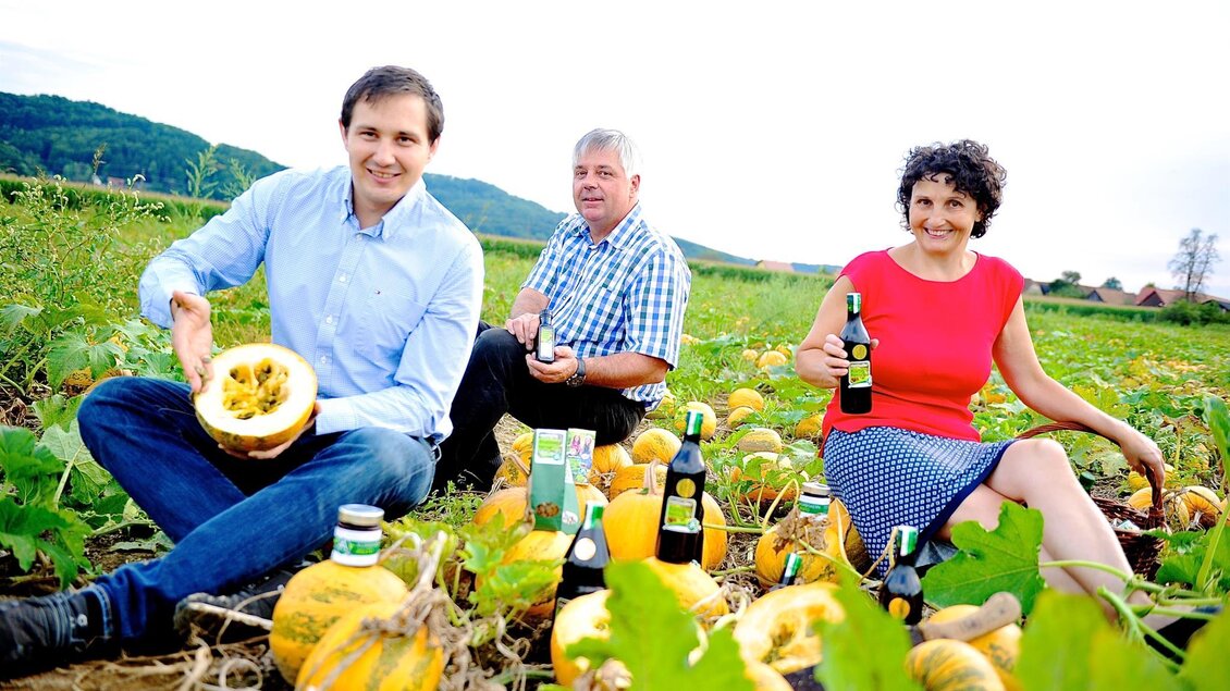 Drei Personen sitzen in einem Kürfield und halten Kürbisse und Getränke in den Händen. Die Landschaft ist grün und hügelig im Hintergrund. | © Familie Reiter-Haas