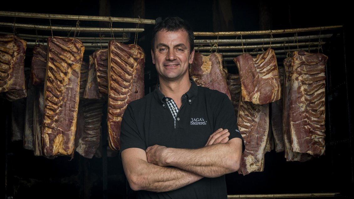 Ein Mann steht stolz mit verschränkten Armen vor einer Auswahl von geräuchertem Fleisch. Die Atmosphäre ist rustikal und traditionell. | © LK Stmk