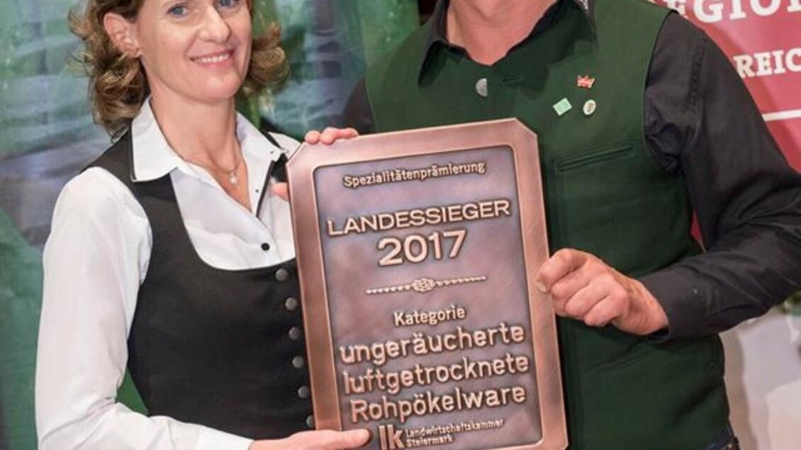 Eine Frau und ein Mann stehen lächelnd zusammen und halten eine Trophäe mit der Aufschrift "Landessieger 2017". Im Hintergrund sind Banner von regionalen Produkten zu sehen. | © LK Stmk