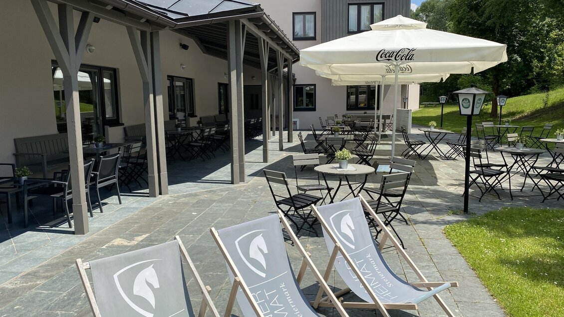Terrasse im JUFA Hotel Maria Lankowitz mit Liegestühlen, Tischen, Sonnenschirmen und Sitzbereich neben dem Gebäude.