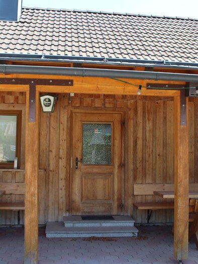 A traditional wooden house with a solid roof. In front of the house, there are wooden benches and tables. | © Jan`s Jausenhütt`n