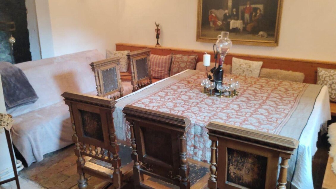 <p>Eine gemütliche und rustikale Essecke mit Holzstisch und Holzstühlen. Am Boden liegt ein warmer Teppich und an der Wand hängt ein altes Gemälde. Der Tisch ist gedeckt mit einer weißen Tischdecke und einer Kerze.</</p> | © Podenhaus