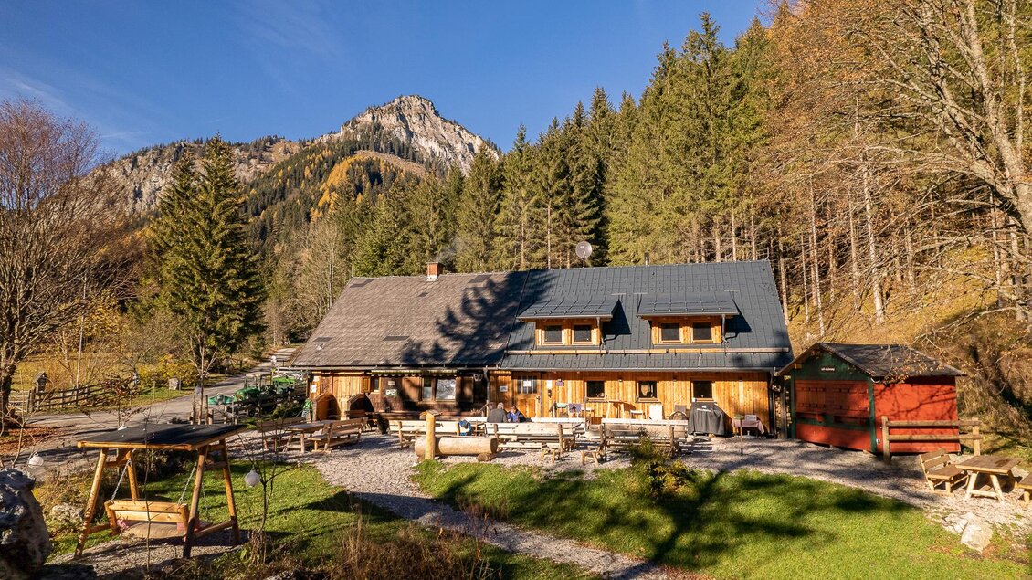 Eine idyllische Hütte umgeben von Bäumen und Bergen. Der Garten ist einladend mit Sitzmöglichkeiten im Freien. | © Jassingalm
