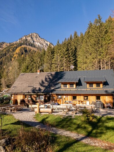 Eine idyllische Hütte umgeben von Bäumen und Bergen. Der Garten ist einladend mit Sitzmöglichkeiten im Freien. | © Jassingalm