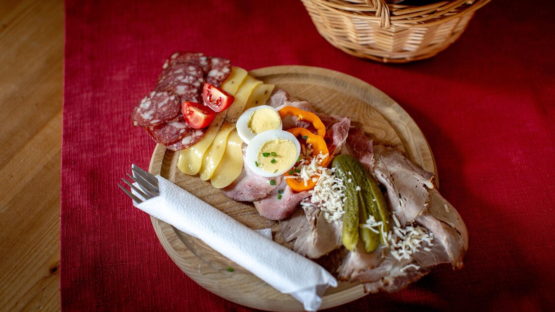 Ein Holzbrett mit einer Auswahl an Wurst, Schinken, Käse und Gemüse. Daneben liegt Besteck und ein Korb mit Brot im Hintergrund. | © Tourismusverband Murau