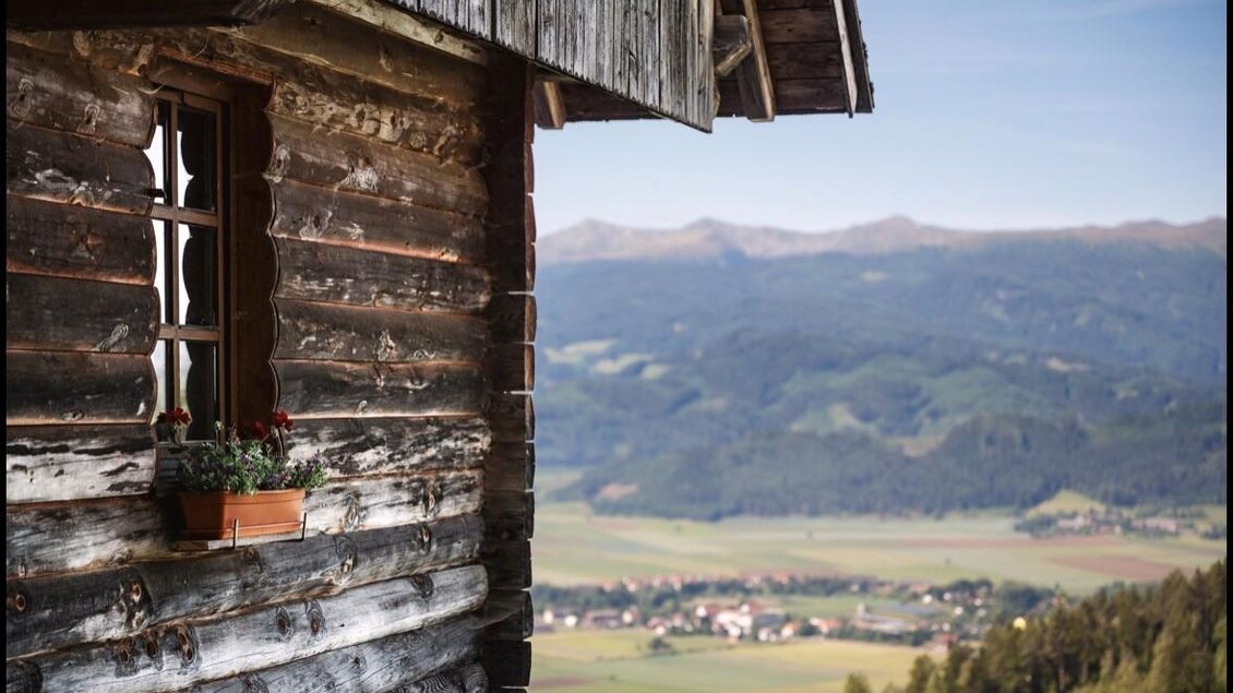 Eine Holzfassade mit einem Fenster und einem Blumenkasten. Im Hintergrund erstreckt sich eine grüne Landschaft mit Hügeln und Dörfern. | © Jausenstation Höhn
