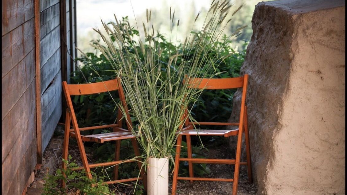 Zwei Holzstühle stehen nebeneinander, dekoriert mit einer großen Vase voller Pflanzen. Im Hintergrund sind sanfte Hügel und ein klarer Himmel sichtbar. | © Jausenstation Höhn