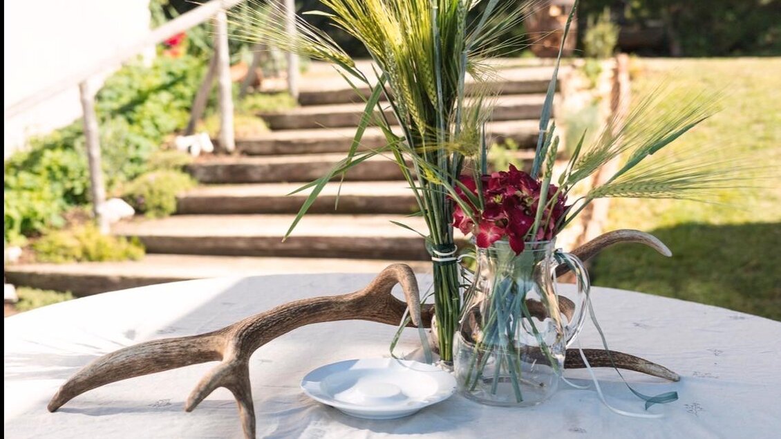 Ein Tisch im Freien, dekoriert mit einer Vase aus Blumen und Pflanzen. Im Hintergrund sind Treppen und grüne Wiesen zu sehen. | © Jausenstation Höhn