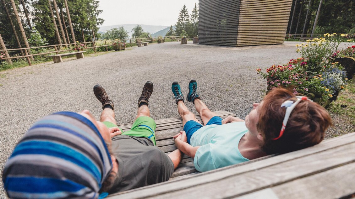 Zwei Personen liegen entspannt auf einer Holzbänke und genießen die Aussicht. Es ist ein ruhiger Ort mit Bäumen und einem klaren Himmel im Hintergrund. | © TV Hochsteiermark, nicoleseiser.at