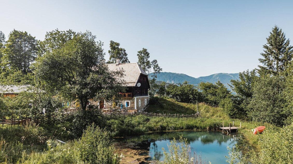 Ein idyllisches Haus inmitten von Bäumen und Wiesen. Neben dem Haus befindet sich ein ruhiger Teich mit Bergen im Hintergrund.