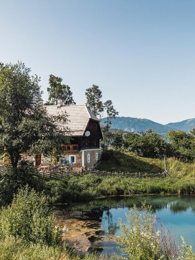 Ein idyllisches Haus inmitten von Bäumen und Wiesen. Neben dem Haus befindet sich ein ruhiger Teich mit Bergen im Hintergrund.