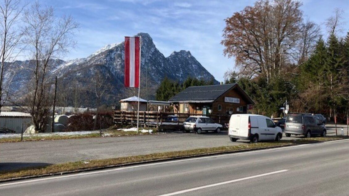 Ein Gebäude mit einer österreichischen Fahne in der Nähe einer Straße, umgeben von Bergen und Bäumen. Die Szenerie zeigt eine ruhige, ländliche Atmosphäre. | © TVB Ausseerland Salzkammergut