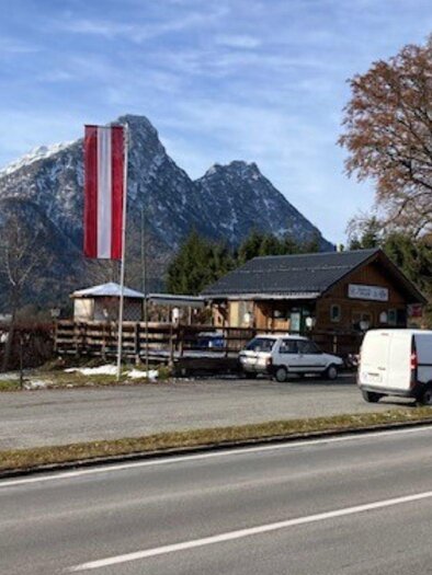 Ein Gebäude mit einer österreichischen Fahne in der Nähe einer Straße, umgeben von Bergen und Bäumen. Die Szenerie zeigt eine ruhige, ländliche Atmosphäre. | © TVB Ausseerland Salzkammergut