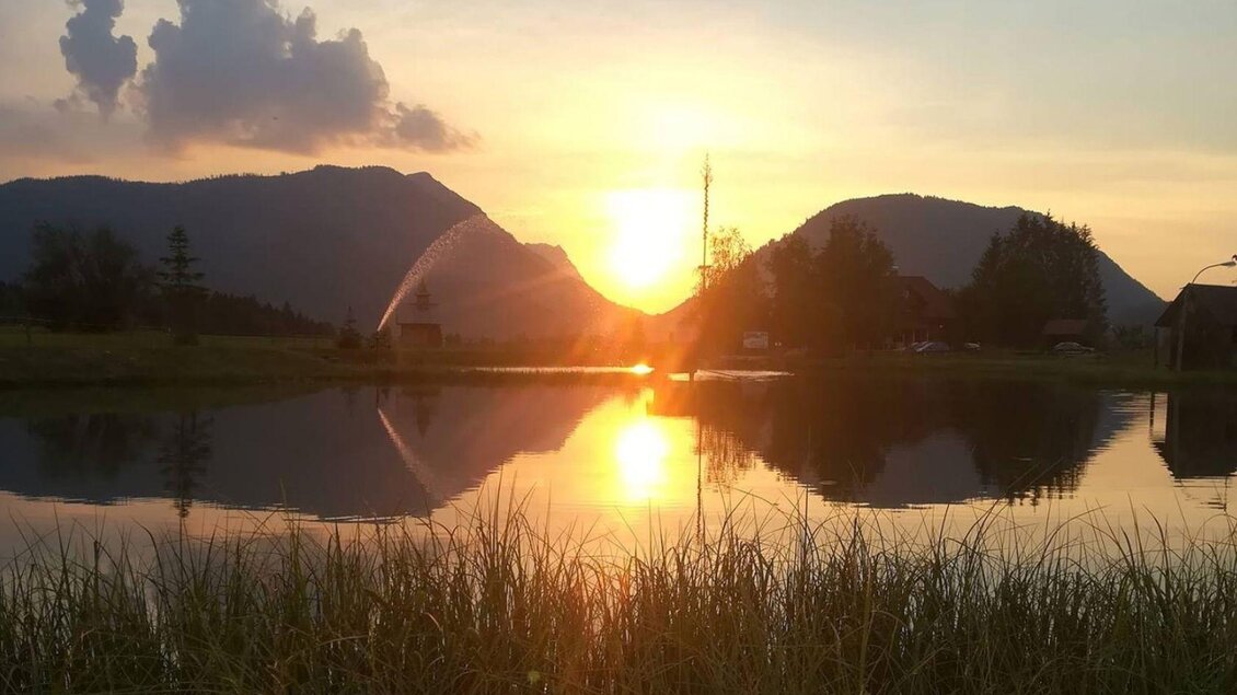 Ein wunderschöner Sonnenuntergang über einem ruhigen See, umgeben von Bergen. Die Sonne spiegelt sich im Wasser und schafft eine malerische Atmosphäre.