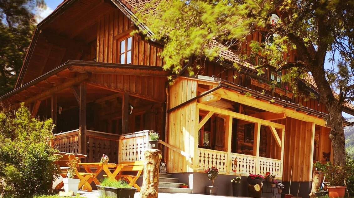 Ein charmantes Holzhaus mit einer großen Veranda und einem einladenden Garten. Umgeben von Bäumen und grüner Wiese ist es ein perfekter Ort zum Entspannen.
