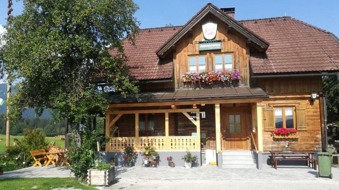 Ein charmantes Holzhaus mit einem Empfangsbereich und bunten Blumenkästen. Im Hintergrund sind grüne Wiesen und ein klarer Himmel zu sehen.