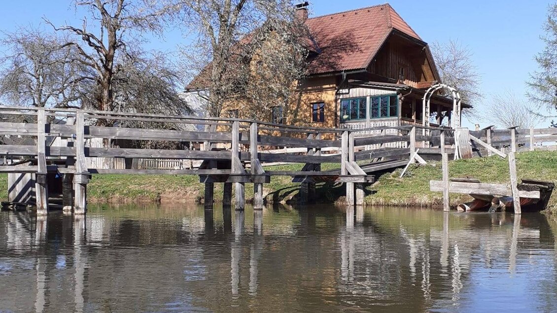 Ein malerisches Haus am Wasser mit einer Holzbrücke. Im Hintergrund sind blühende Bäume und ein klarer blauer Himmel zu sehen.