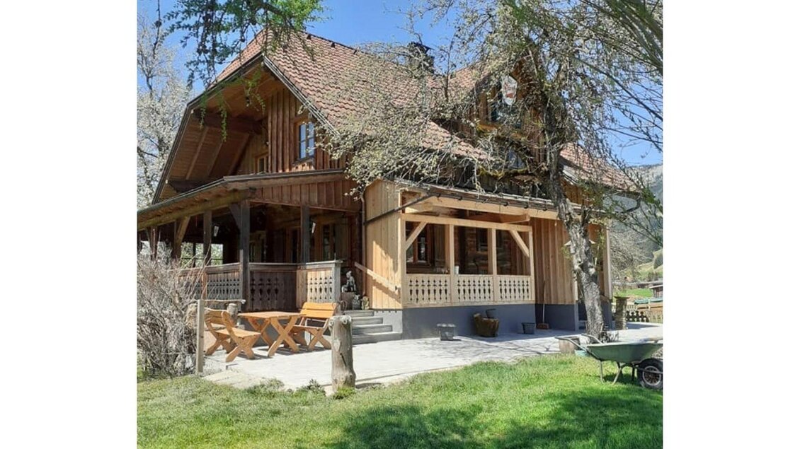 Ein charmantes Holzhaus mit einem großen Balkon. Der Garten ist grün und einladend, umgeben von Bäumen.