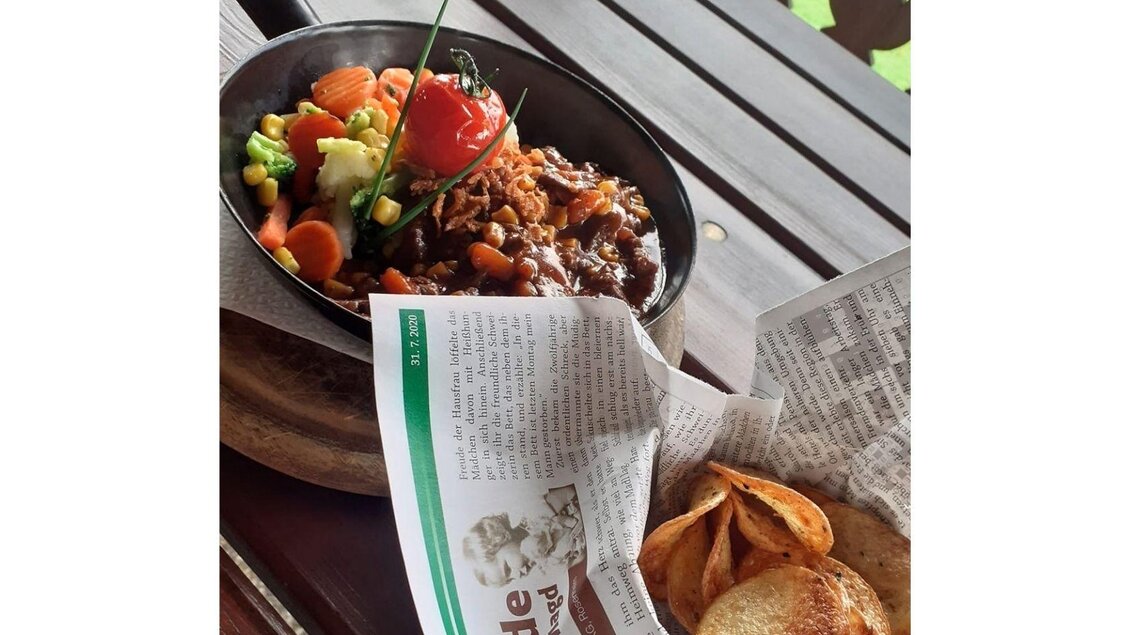 Ein köstliches Gericht in einer Pfanne mit Gemüse und einer Beilage aus Kartoffelchips. Das Essen ist auf einer Holztischplatte angerichtet, umgeben von einer Zeitungsunterlage.