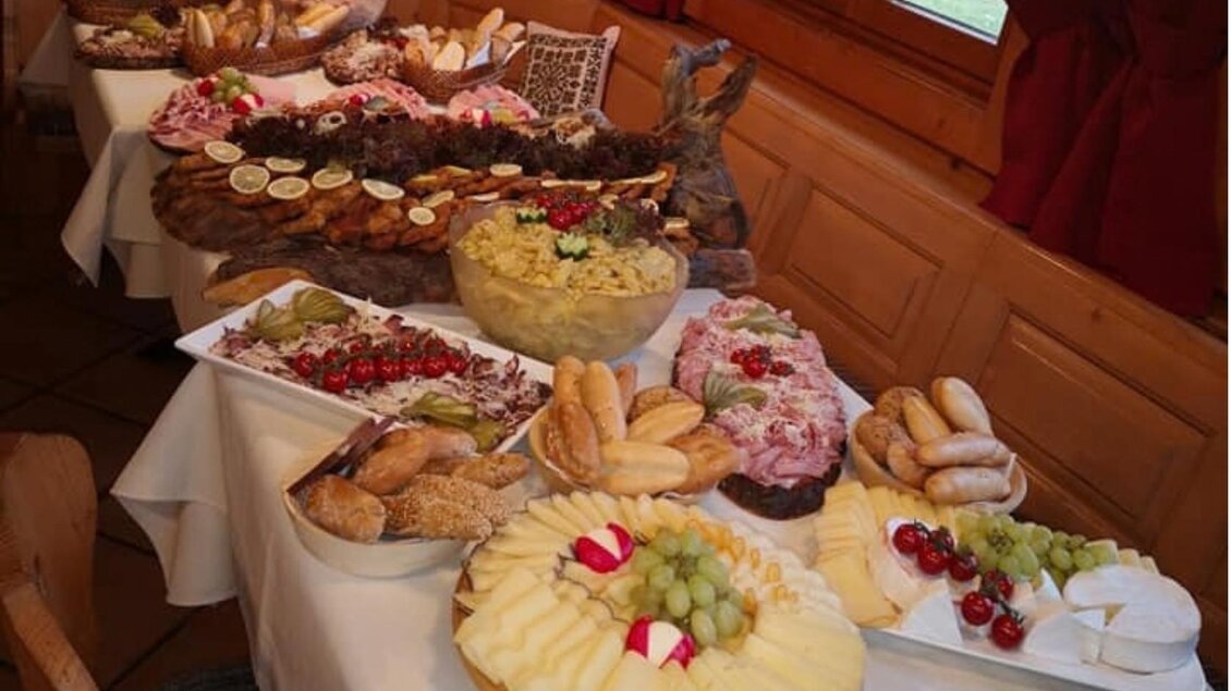 Ein reichhaltiges Büfett mit verschiedenen Leckereien, darunter Kuchen, Obst und Brötchen. Der Tisch ist in einem gemütlichen Raum mit Holzverkleidung gedeckt.