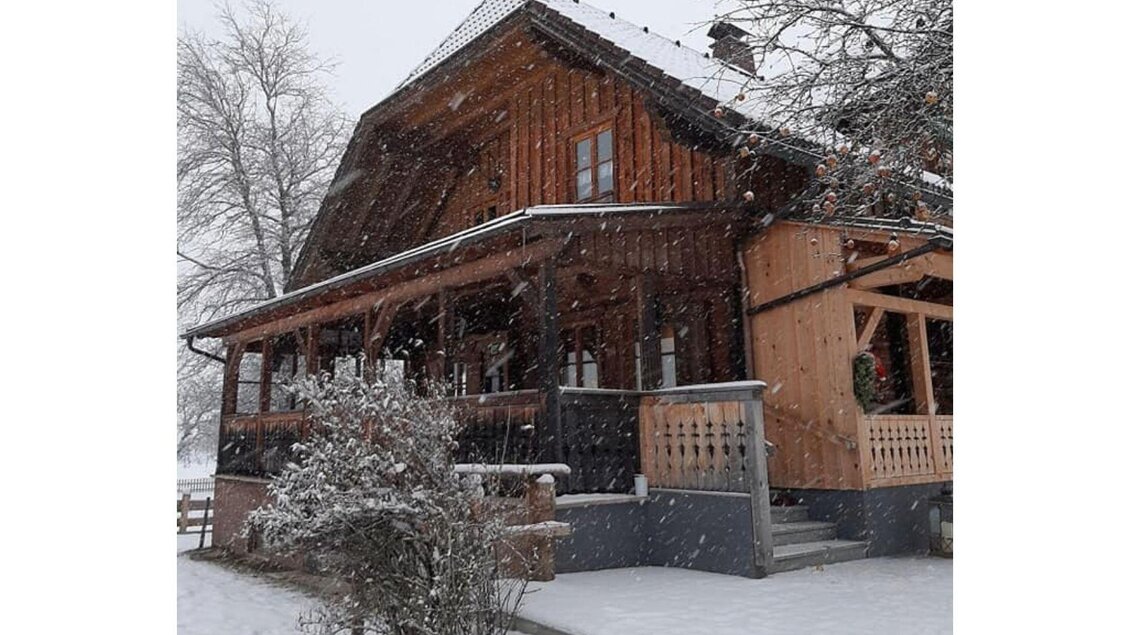 Ein charmantes Holzhaus im Schnee, umgeben von einem winterlichen Landschaftsbild. Großer Schnee fällt auf das Dach und den Vordereingang.