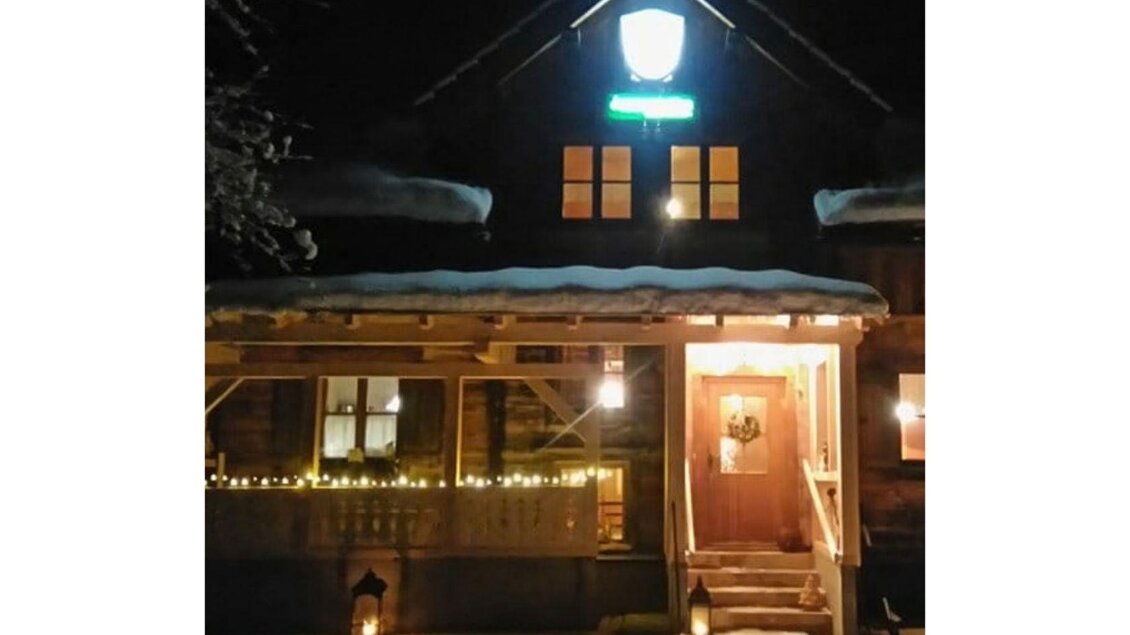 Ein gemütliches Holzhaus bei Nacht, beleuchtet von warmem Licht. Schneebedeckter Boden und festliche Dekorationen schaffen eine einladende Atmosphäre.