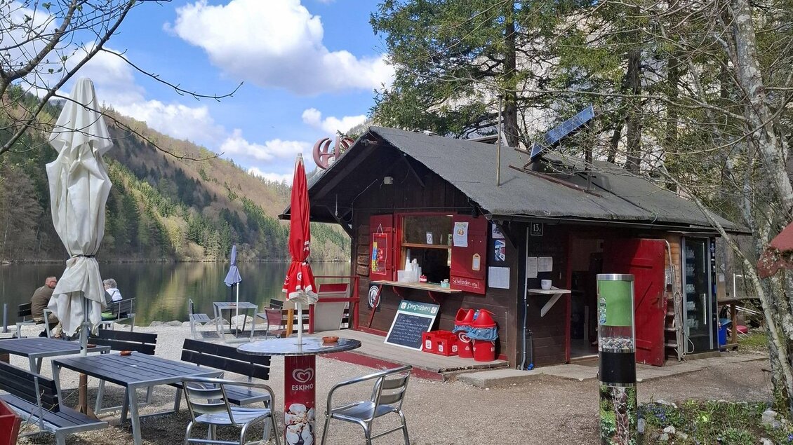 Eine gemütliche Hütte in der Natur, umgeben von Bäumen. Menschen sitzen entspannt auf Bänken und genießen die Umgebung. | © Jausenstation am Leopoldsteinersee