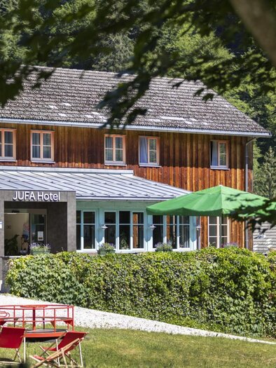 Ein gemütliches Holzhaus mit einem einladenden Eingangsbereich. Umgeben von grünen Büschen und Sonnenschirmen im Freien. | © Jufa Hotel Eisenerz