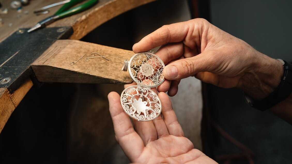 Hände halten ein geöffnetes filigranes Schmuckstück aus Metall zur Reparatur an der Werkbank von Juwelier Grassl. | © Bernd Grosseck
