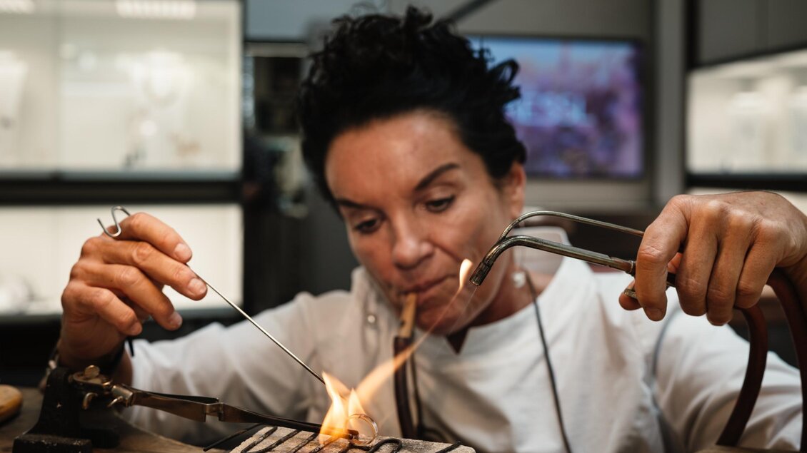 Frau Gressl bearbeitet ein neues Schmuckstück mit einer Flamme und feinem Werkzeug an der Werkbank. | © Bernd Grosseck