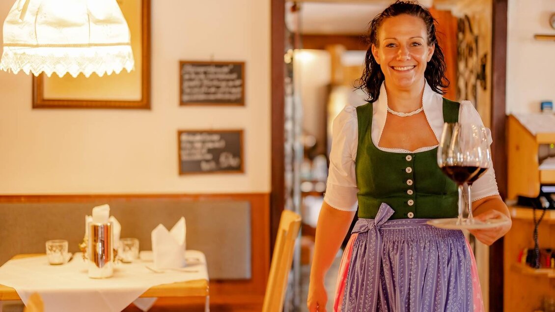 Eine lächelnde Kellnerin trägt ein Glas Wein in einem gemütlichen Restaurant. Die Einrichtung ist rustikal und einladend. | © K.U.K. Wirtshaus