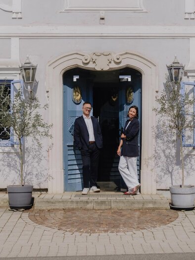 Ein elegantes Gebäude mit blauen Fensterläden und zwei Personen, die vor der Tür stehen. Der Boden ist gepflastert und es gibt einige Topfpflanzen. | © Michaela Lorber