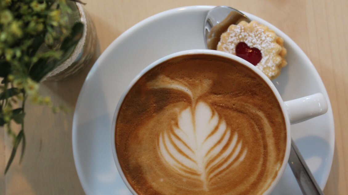 Eine Tasse Kaffee mit einer schönen Latte Art und einem Keks auf einem weißen Teller. Daneben steht ein kleiner Topf mit einer grünen Pflanze. | © Katharina Zeiler