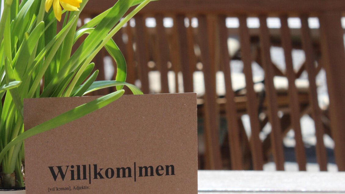 Ein Willkommensschild steht auf einem Holztisch, umgeben von gelben Blumen in einem Topf. Im Hintergrund sind Holzstühle und eine grüne, strukturierte Wand sichtbar. | © Katharina Zeiler