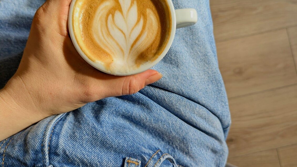 Eine Hand hält eine Tasse Kaffee mit Latte Art. Die Person sitzt auf einem Stuhl und trägt eine schwarze T-Shirt und Jeans. | © Zeiler Katharina